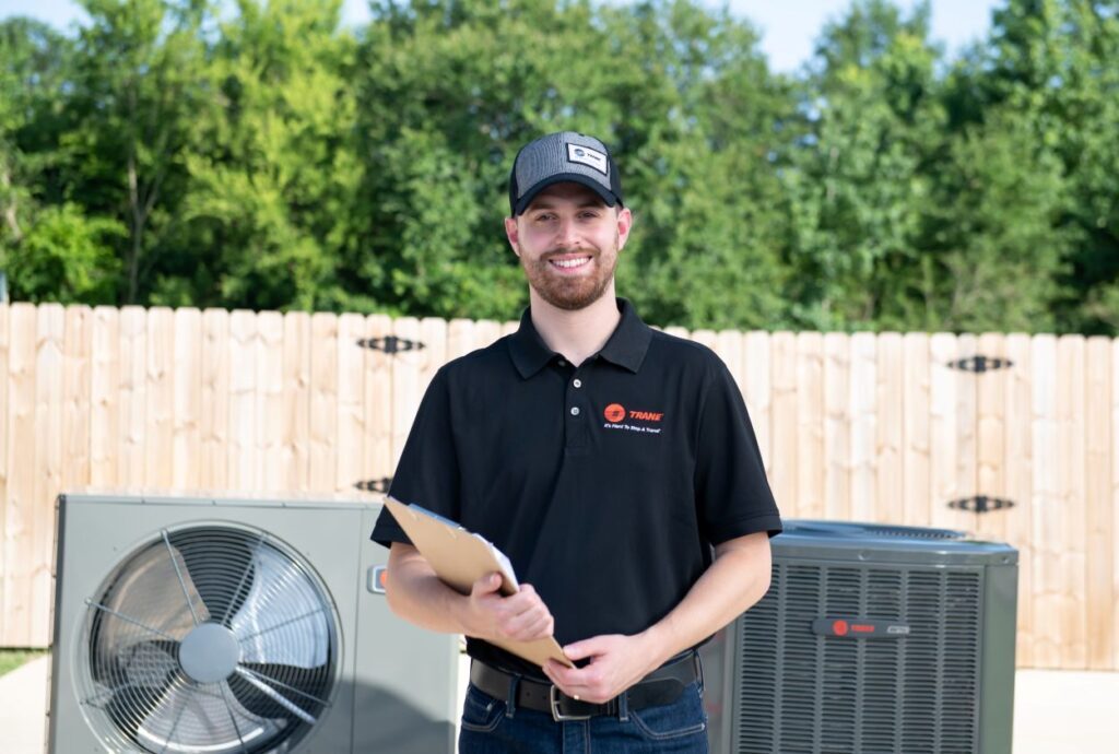 Smiling HVAC technician in a Trane uniform holding a clipboard, standing outdoors in front of Trane air conditioning units with a wooden fence and trees in the background
