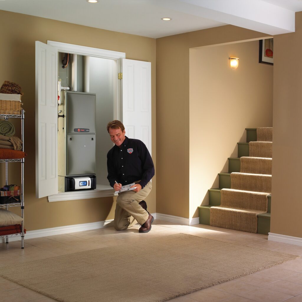 HVAC technician kneeling in a finished basement while inspecting a Trane furnace system installed in a wall closet, taking notes on a clipboard