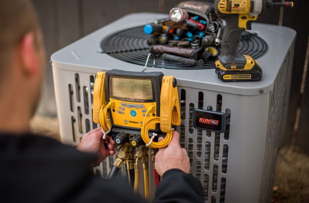 HVAC technician using a Fieldpiece digital manifold gauge to service a RunTru air conditioning unit with tools and a cordless drill placed on top