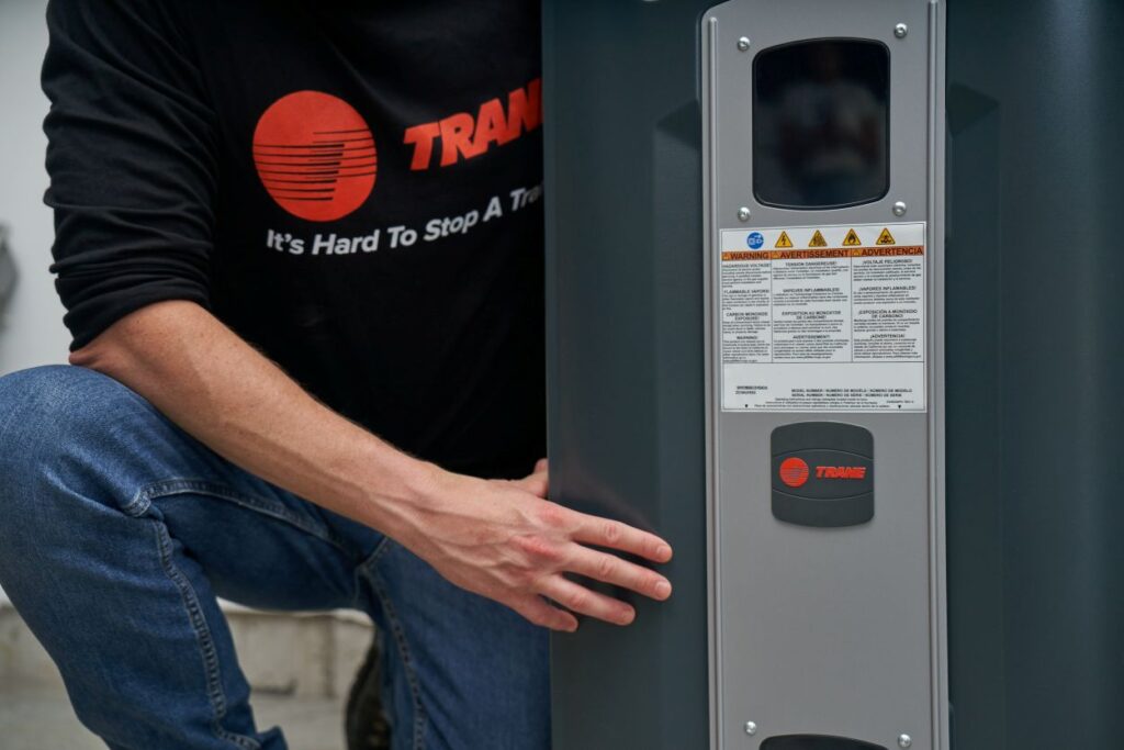 HVAC technician in a Trane shirt inspecting a Trane heating and cooling unit with warning labels and branding visible on the side panel